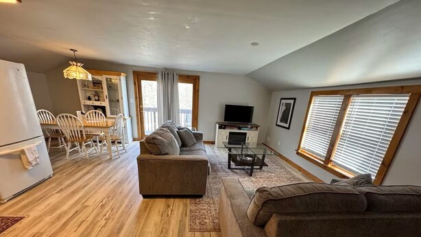 Un espacio habitable amplio con piso de madera, un conjunto de sofás, una mesa de comedor con sillas, un televisor y una ventana con persianas.