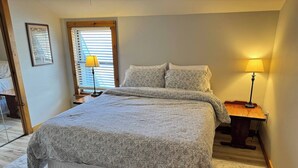 Un dormitorio con una cama grande, dos lámparas de noche, una ventana con persianas y suelo de madera.