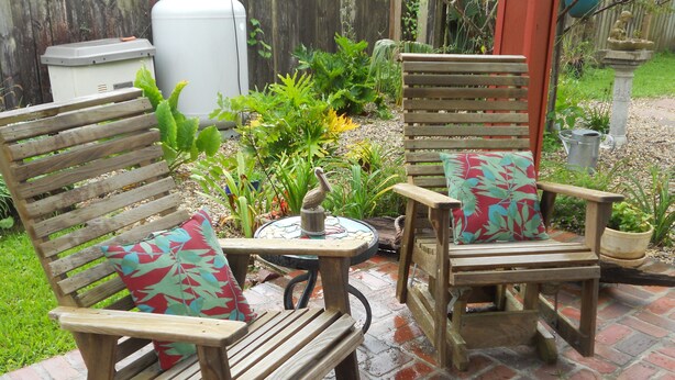 Two wooden rocking chairs with colorful cushions, a small table with a birdbath, and a garden with various plants and a fence in the background. - Cozy Living Close to the Beach (Bay St Louis)