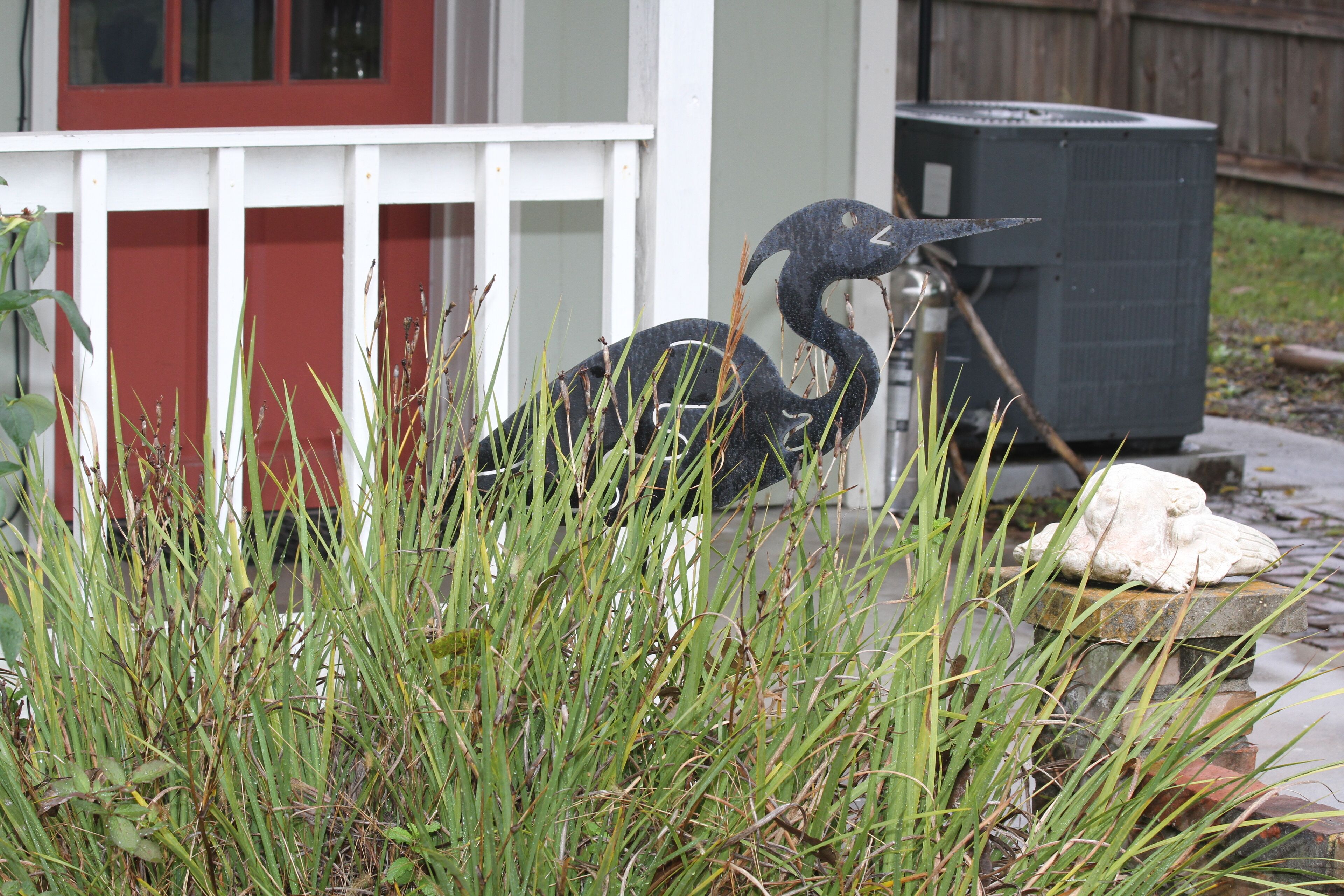 Une sculpture d’oiseau, posée sur un piédestal en pierre, dans un jardin délimité par une clôture blanche, avec une porte rouge en arrière-plan.