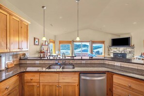 A kitchen with wooden cabinets, a stainless steel dishwasher, a granite countertop, pendant lights, a stone fireplace, and a large window with a view.