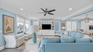 A spacious living room with a large blue sofa, a ceiling fan, and a flat-screen TV. - 🌊Less Stress: Oceanfront Home with Private Pool + Elevator🌊 (Kure Beach)