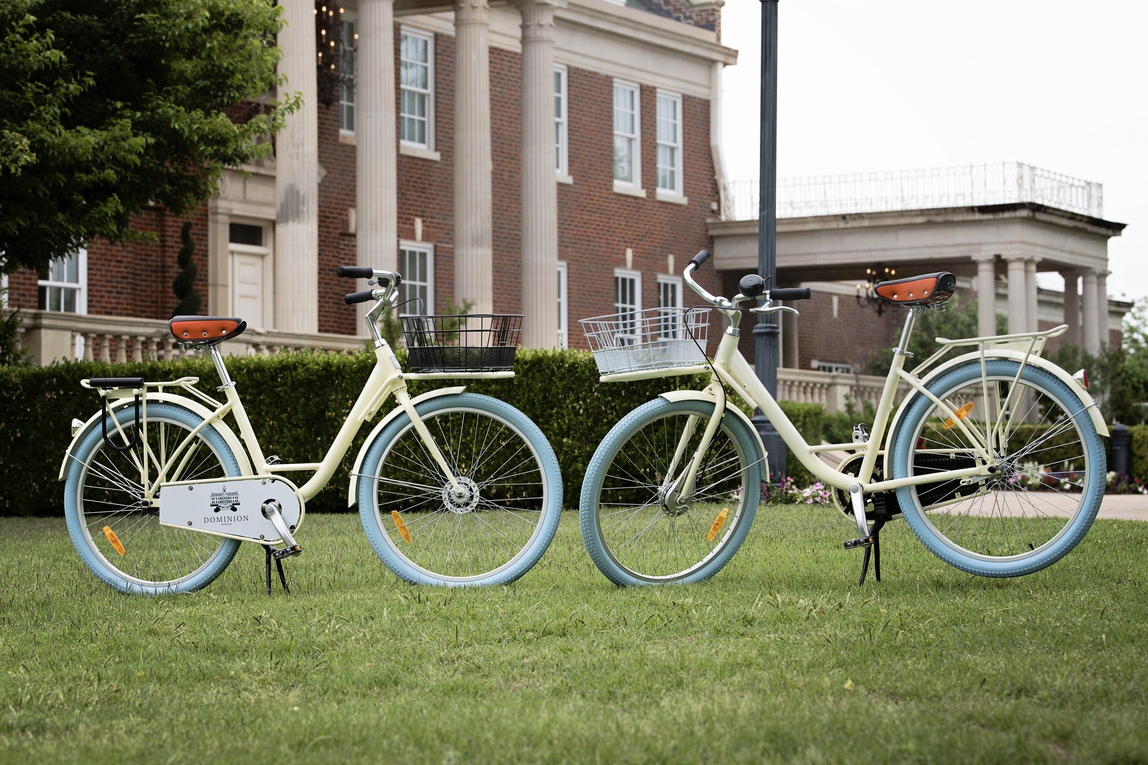 Two vintage bicycles parked on grass with a brick building and a bridge in the background.