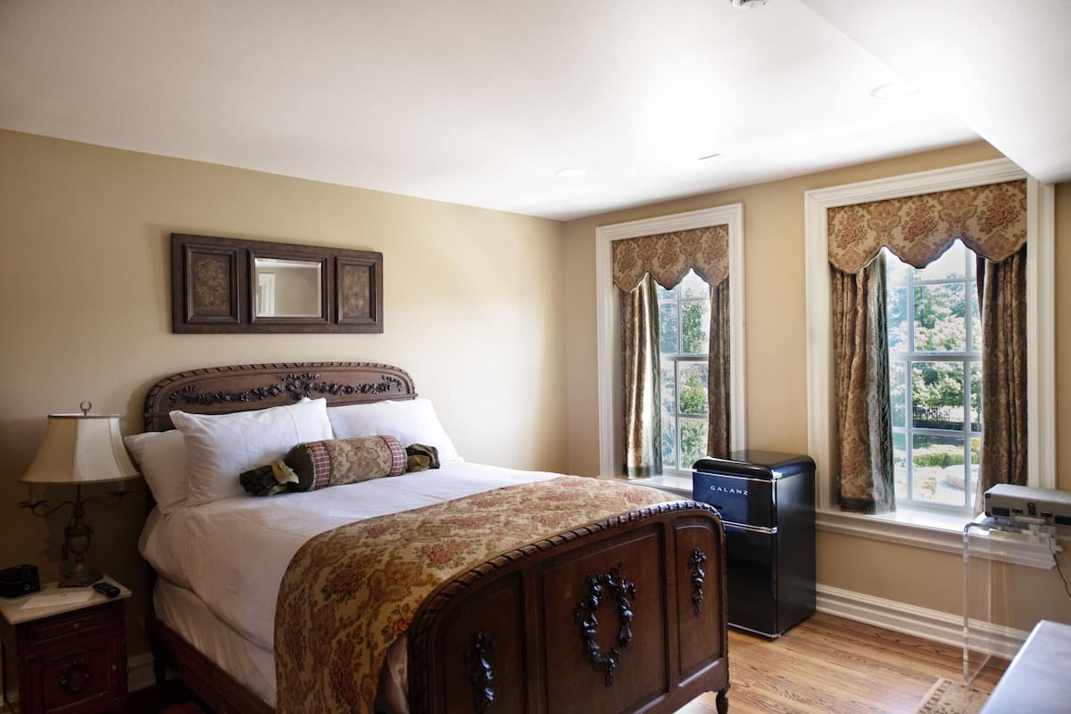 A bedroom with a large bed, a bedside table with a lamp, a dresser, and a window with curtains.