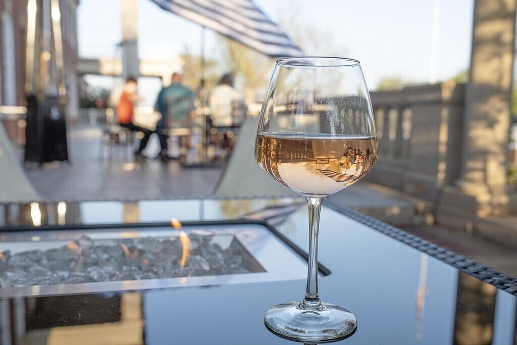 Ein Glas Weißwein auf einem Tisch mit Blick auf eine Terrasse und verschwommene Personen im Hintergrund.