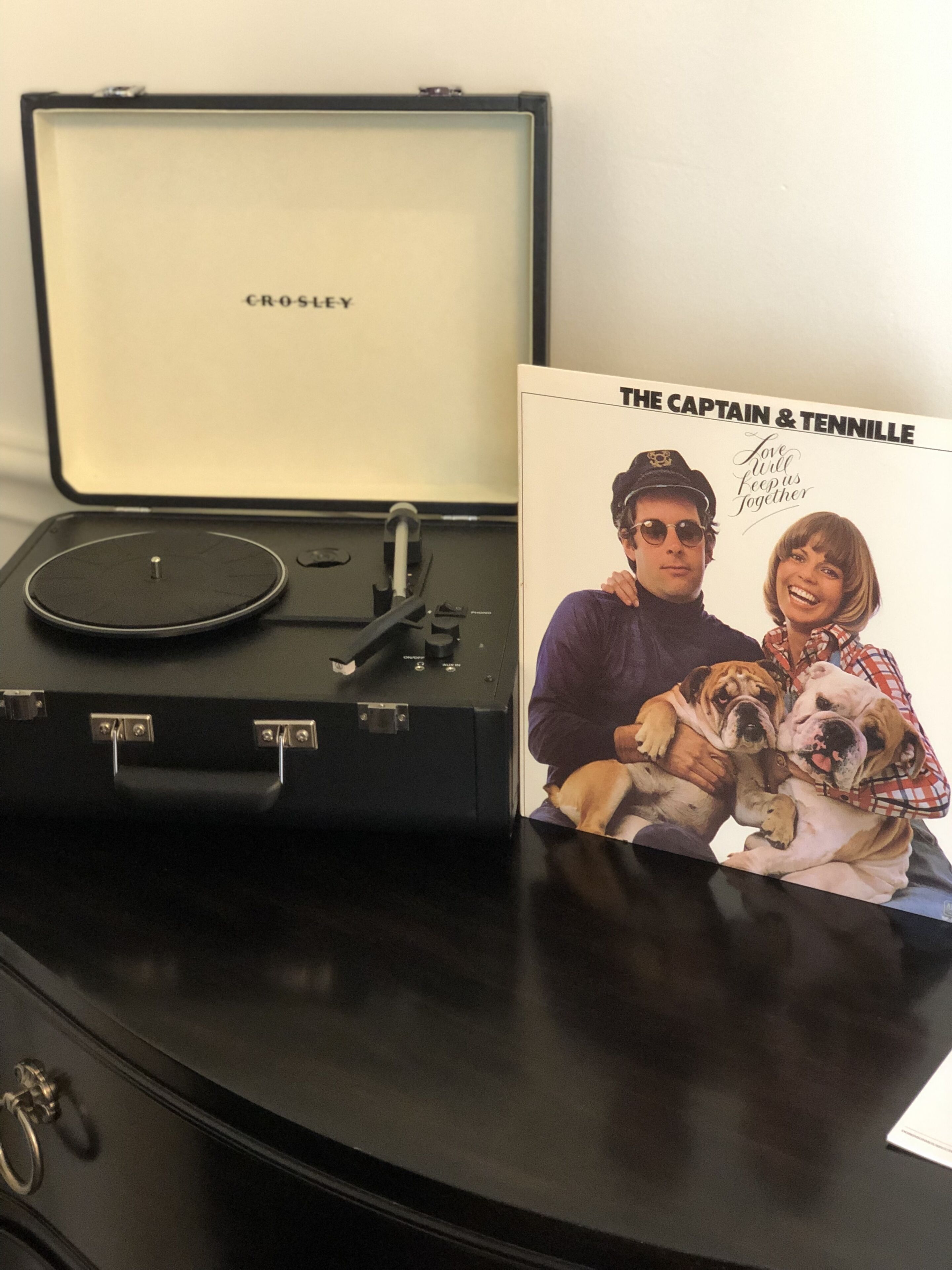 Un giradischi portatile Crosley su una scrivania, con un vinile e la copertina di un CD dei 'The Captain & Tennille' accanto.