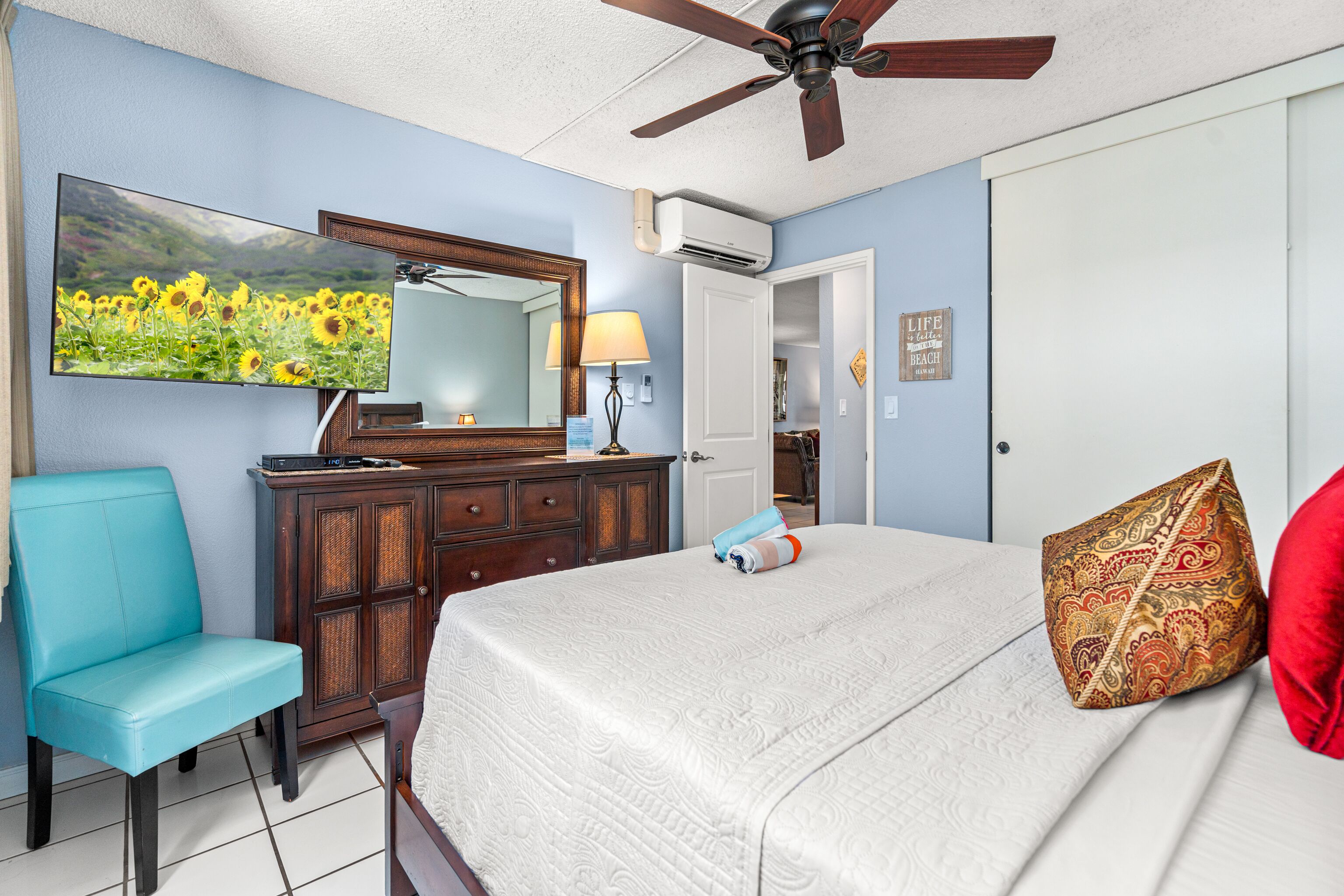 A bedroom with a bed, a ceiling fan, a dresser, a chair, a TV, and a door.
