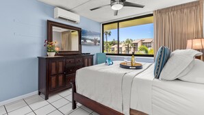Hotelkamer met een bed, een dressoir, een plafondventilator, een raam met uitzicht op palmbomen en gebouwen, en een airconditioner.