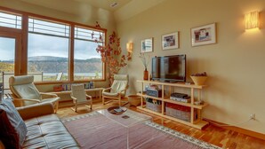 Ein Wohnzimmer mit einer Couch, zwei Sesseln, einem Couchtisch, einem Fernseher und einem Bücherregal. Große Fenster bieten einen Blick ins Freie.