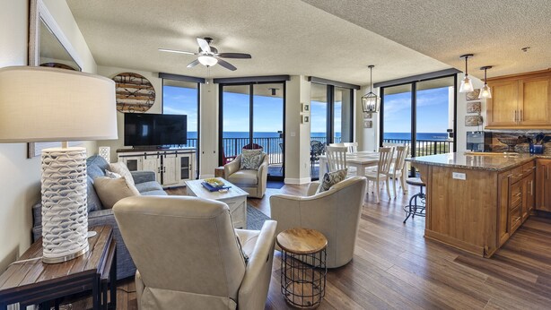 A spacious living area with a large window, a ceiling fan, and a kitchen with wooden cabinets. - Phoenix 8 1101 | Beachfront View | Spacious Corner Condo | My Beach Getaways (Orange Beach)