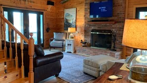 Ein gemütliches Wohnzimmer mit Kamin, einem an der Wand befestigten Fernseher, einer Holzbalkendecke und einer Treppe, die eine weitere Ebene hinaufführt.