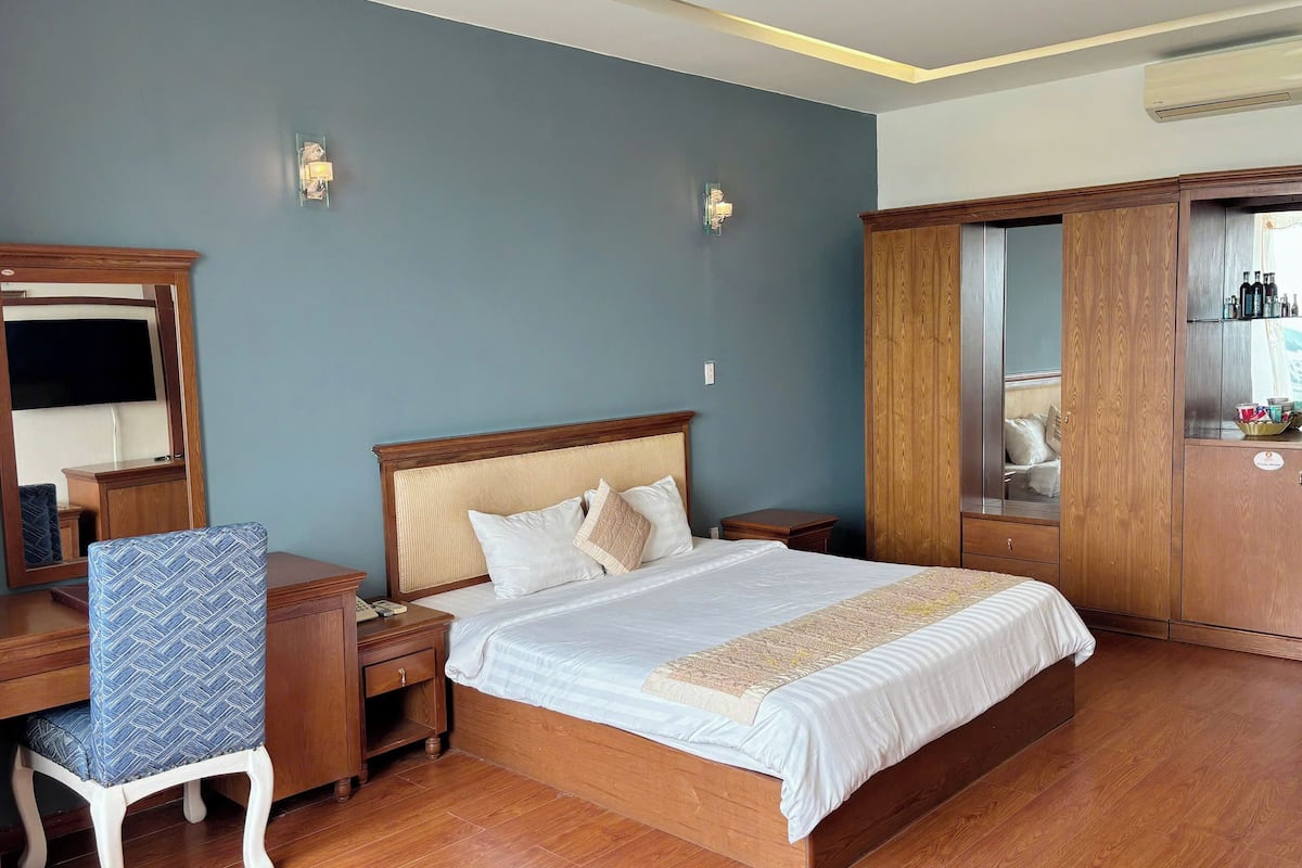 Quarto de hotel com cama de madeira, mesa de cabeceira, cadeira, guarda-roupa e espelho.