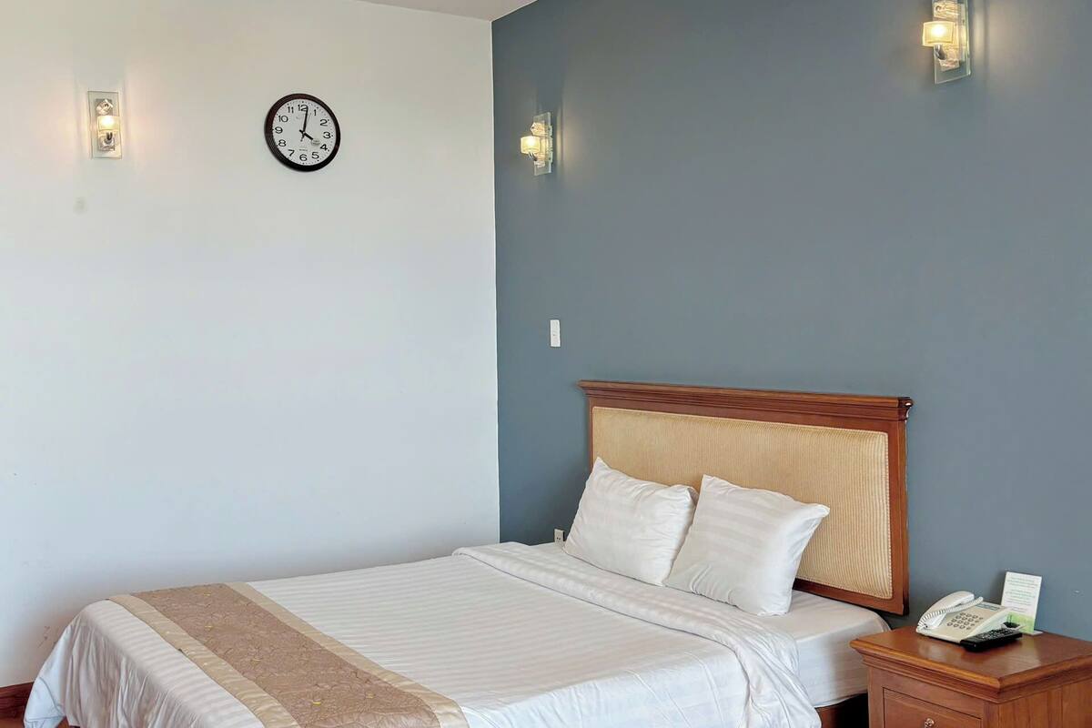 Cama bem arrumada com cabeceira de madeira, mesa de cabeceira com telefone e um letreiro, e um relógio de parede.