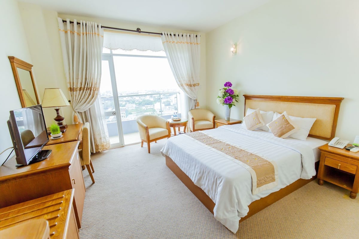 Quarto de hotel com cama, escrivaninha com computador, cadeira, mesinha e criado-mudo com telefone.