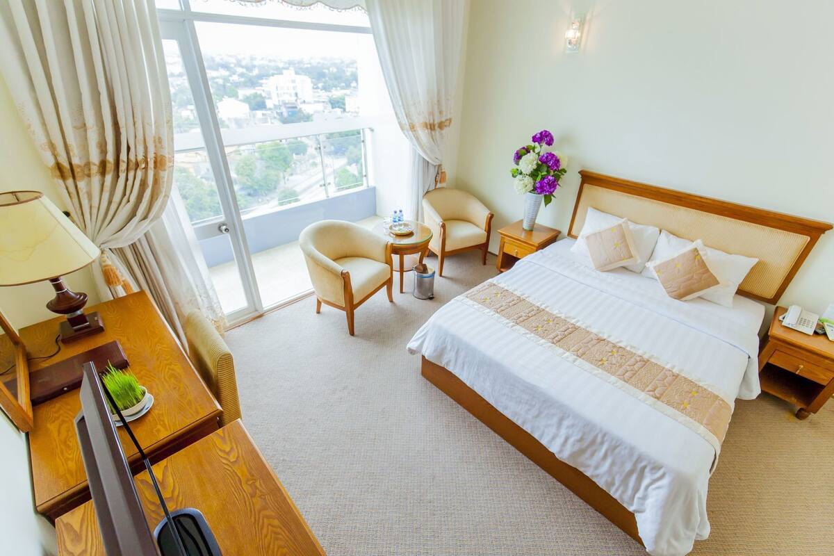 Quarto de hotel com uma cama grande, uma escrivaninha com luminária, uma cadeira, uma pequena mesa com um vaso de flores e vista para a cidade pela janela.