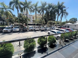Um estacionamento com vários carros e um ônibus, cercado por palmeiras e edifícios.