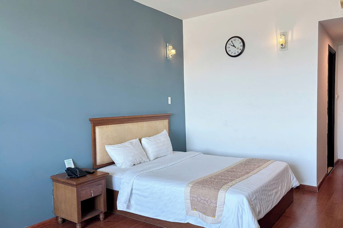 Uma cama bem arrumada com cabeceira de madeira, mesa de cabeceira com abajur e relógio de parede.