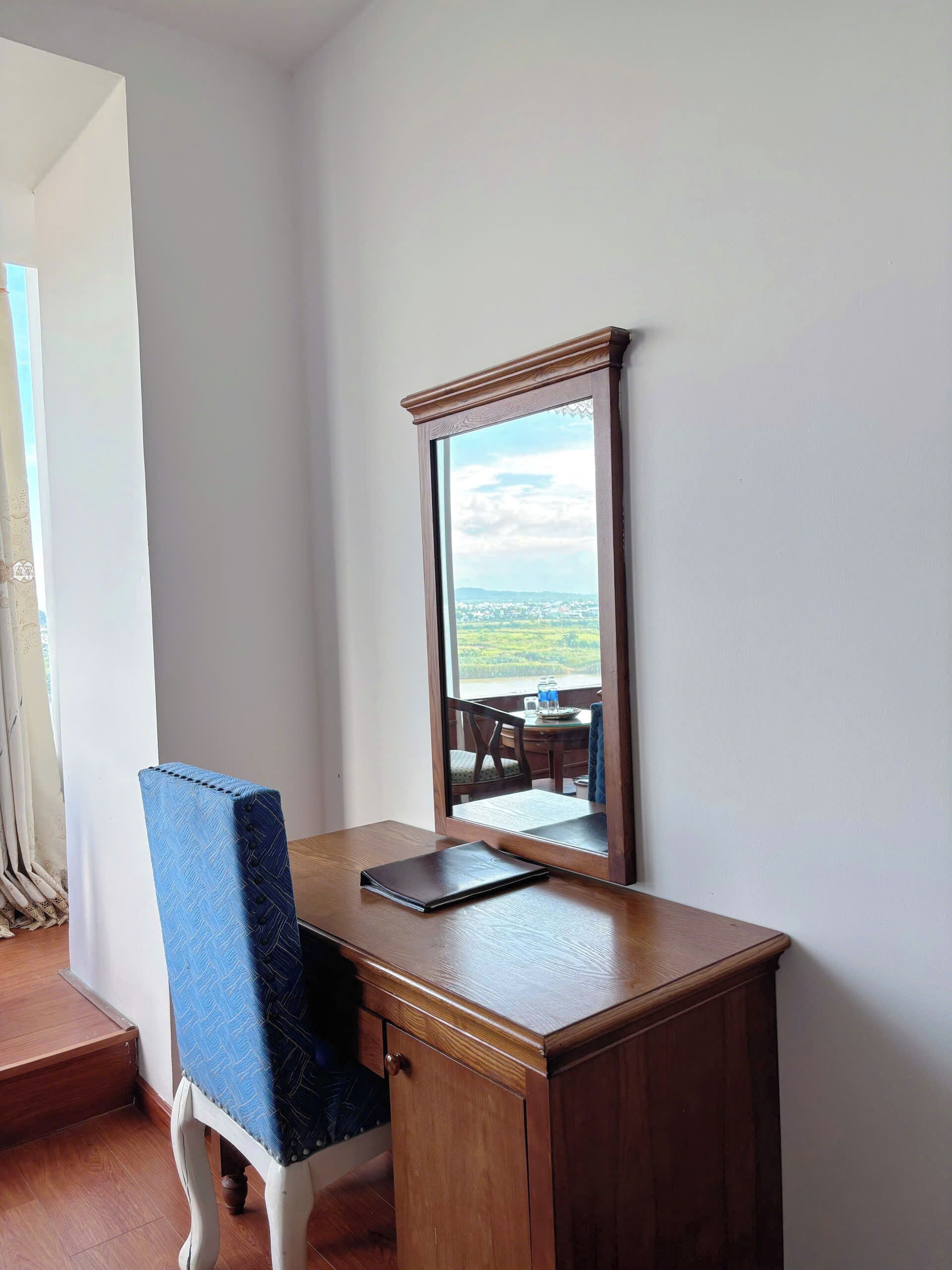 Un escritorio de madera con un espejo, una silla azul y vistas a un paisaje por la ventana.