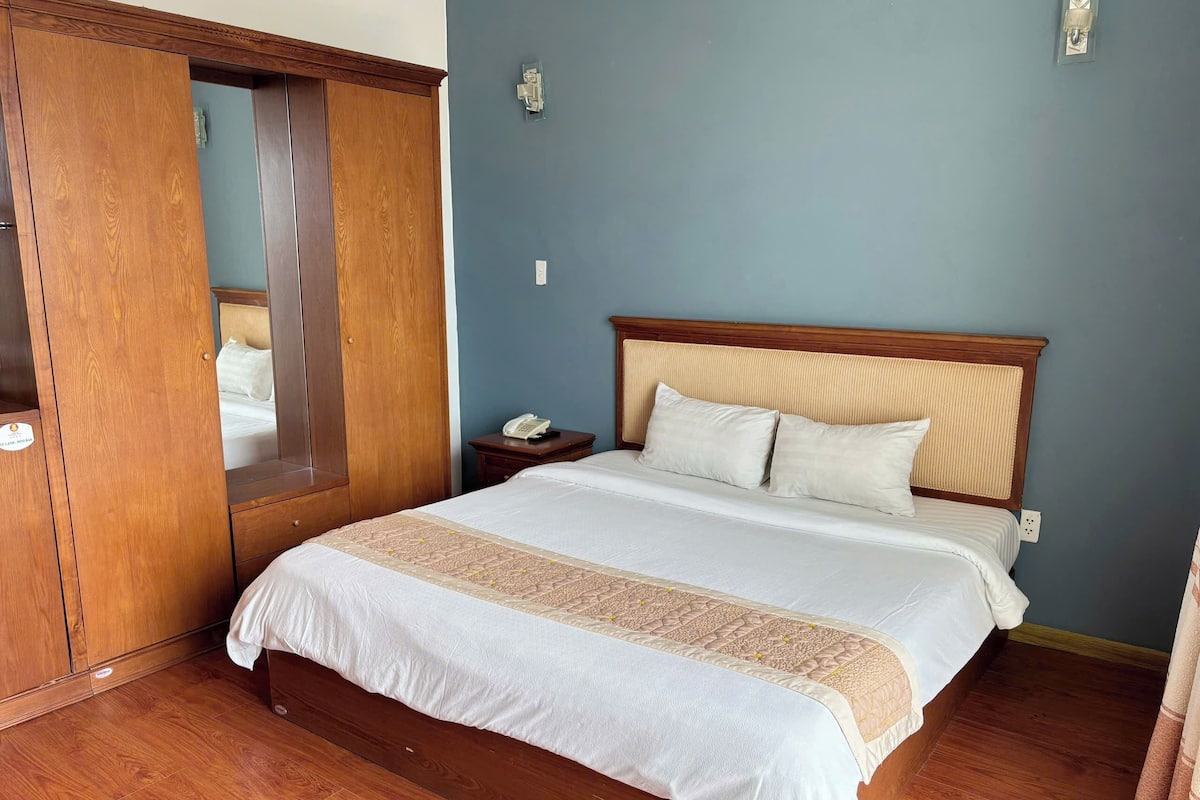 Uma cama bem arrumada com cabeceira de madeira, mesa de cabeceira com telefone e guarda-roupa de madeira com espelho.