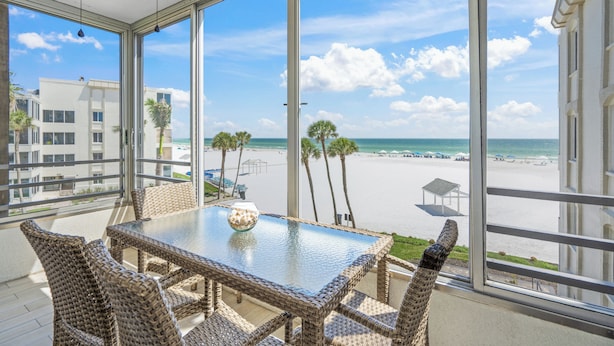 Een balkon met rieten meubels en een glazen tafel, met uitzicht op een strand met palmbomen en een heldere lucht.