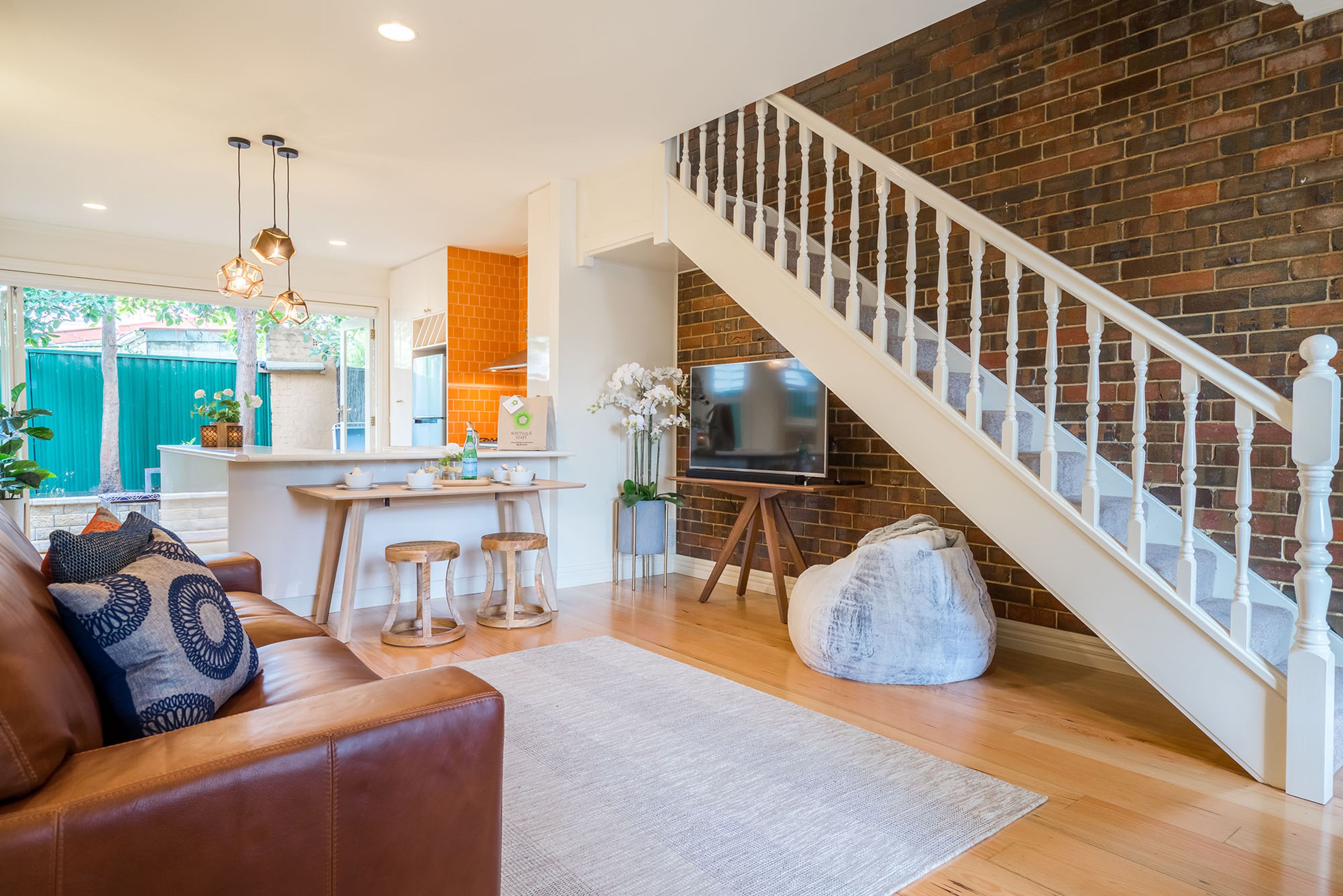 A living room with a brown leather sofa, wooden coffee table, and a white rug. A staircase with white balusters leads to a brick wall.