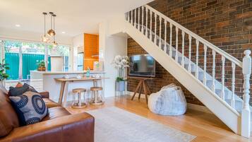 A living room with a brown leather sofa, wooden coffee table, and a white rug. A staircase with white balusters leads to a brick wall.