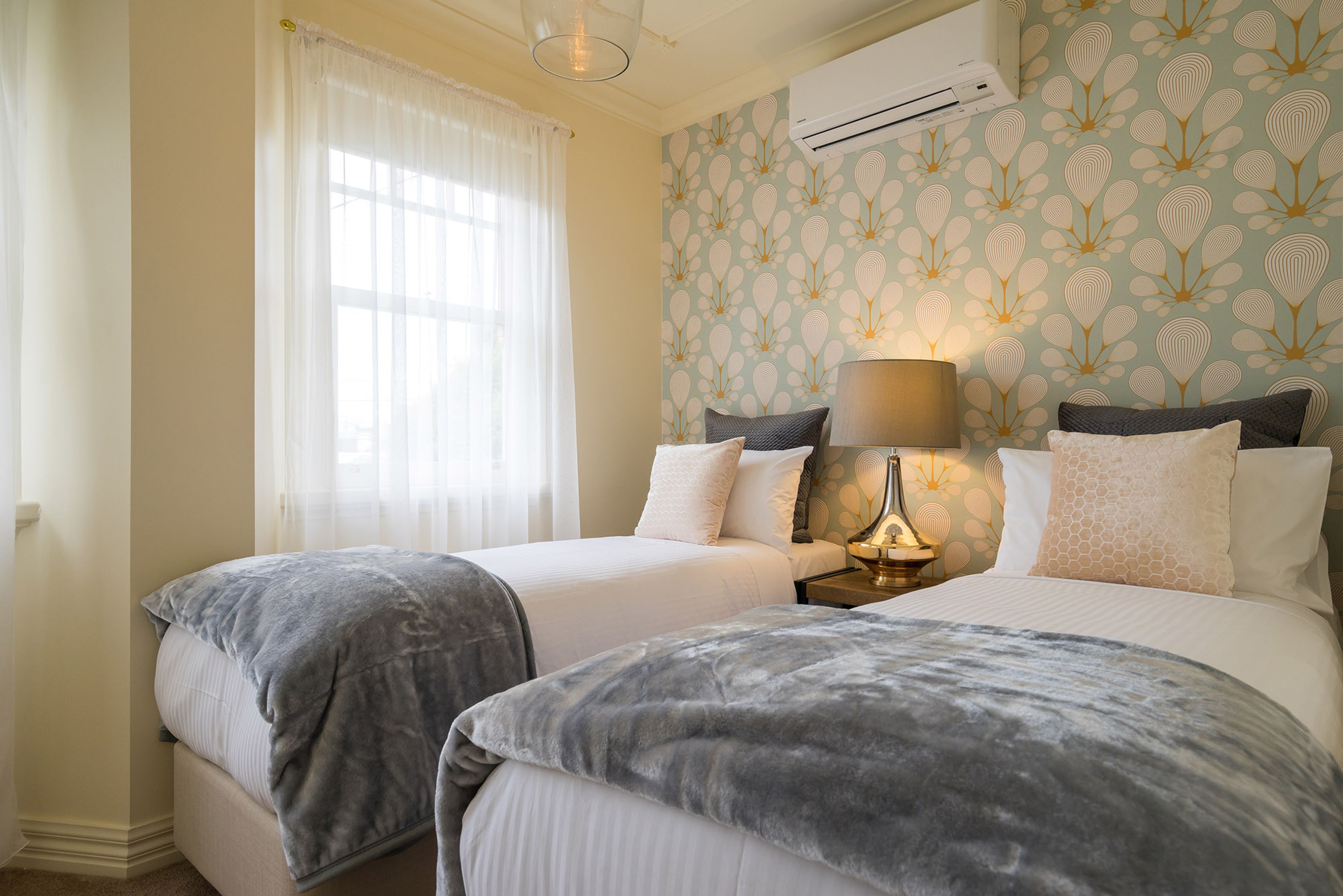 A hotel room with two beds, a nightstand with a lamp, an air conditioning unit, and a window with sheer curtains.