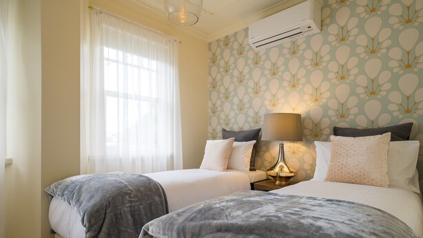 A hotel room with two beds, a nightstand with a lamp, an air conditioning unit, and a window with sheer curtains. - Buckingham Pet friendly (Richmond)