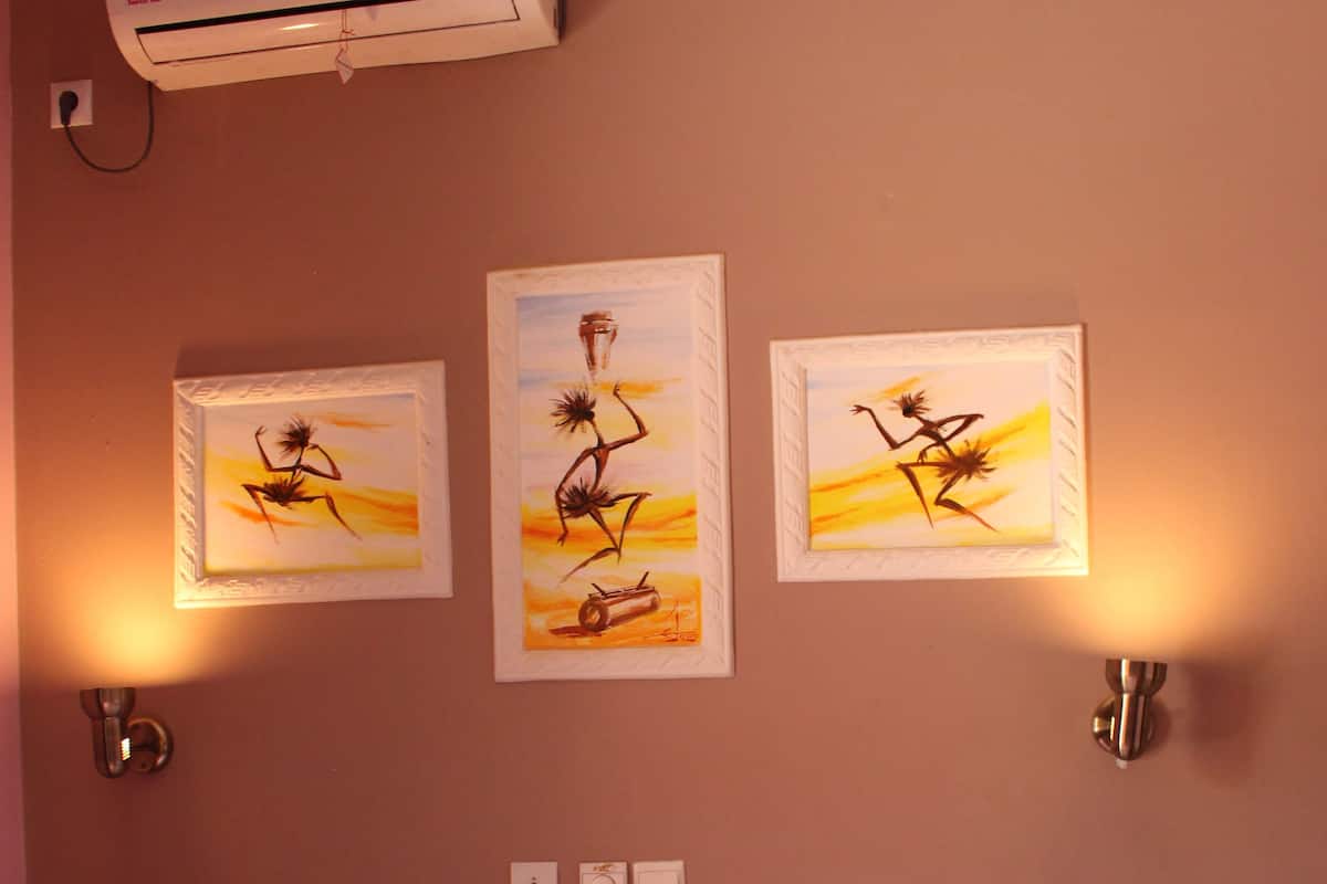 A hotel room with a bed, three wall-mounted artworks, an air conditioner, and a wall-mounted light fixture.