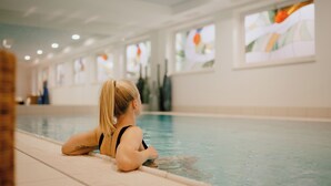 En person sidder ved en indendørs swimmingpool med flisegulv og store vinduer med kunstværker.
