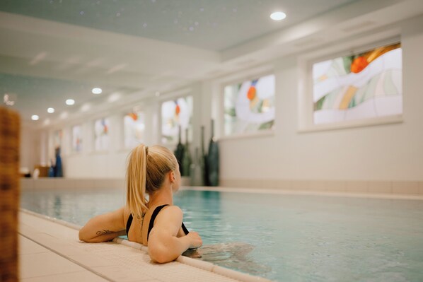 A person sitting by an indoor pool with a tiled floor and large windows with artwork. - Arthotel Gratschwirt (Dobbiaco)