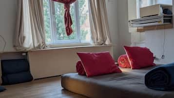 Een slaapkamer met een bed, kussens, een raam met gordijnen, een radiator en een plank met spullen.