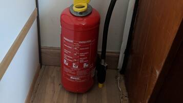 A red fire extinguisher with a yellow handle in a corner of a room with wooden flooring and a white wall.