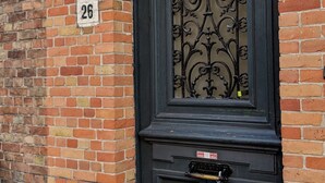 A black metal door with a decorative pattern, a mailbox, and a number '26' sign on a brick wall. - be house (Bruges)
