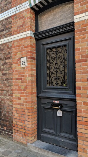 Une porte en métal noir ornée d’un motif décoratif, une boîte aux lettres et une plaque portant le numéro « 26 » sur un mur de briques.