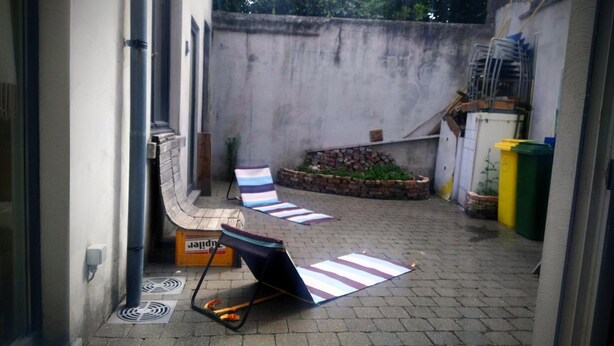 A small outdoor patio area with a brick floor, a metal chair, a striped mat, a small garden bed with plants, and a yellow and green trash bin. - be house (Bruges)