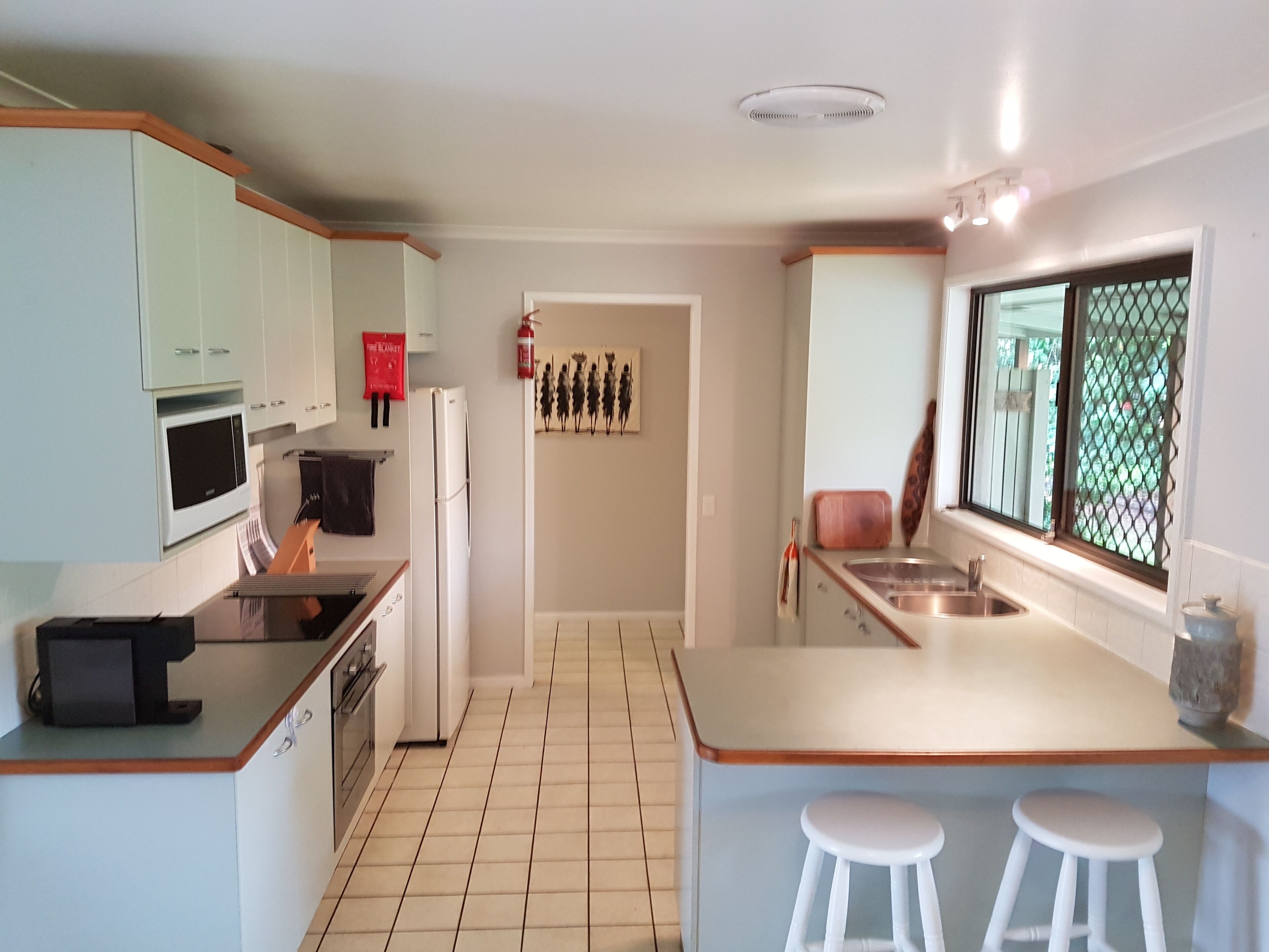 A modern kitchen with white cabinets, a microwave, a refrigerator, and a sink. There is a fire extinguisher on the wall and a window with a view of greenery.