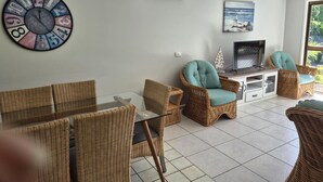 A living room with wicker furniture, a glass table, a wall clock, a television, and a painting of a beach scene. - Beerwah House, 3Bedroom , swimming pool, near Australia Zoo, Sunshine Coast (Beerwah)