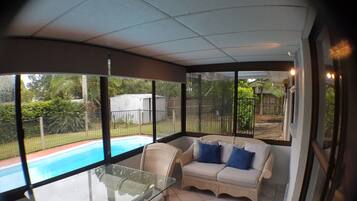 A sunroom with a glass table, wicker sofa, and a view of a backyard swimming pool.