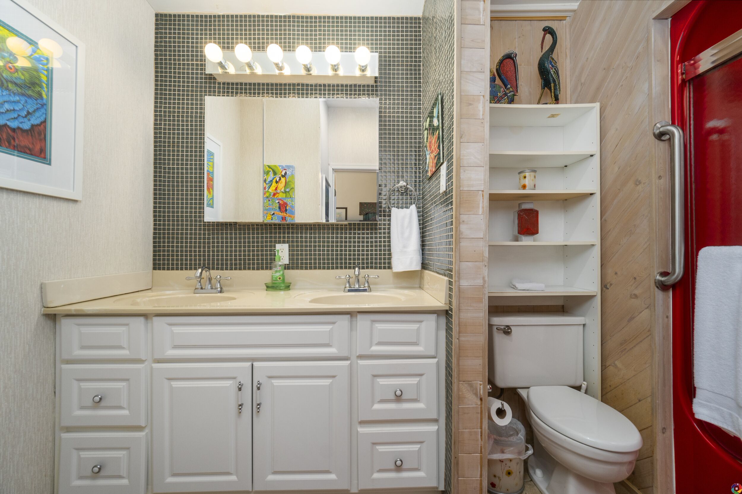 A bathroom with a sink, mirror, and toilet. There are shelves with toiletries and a red door.
