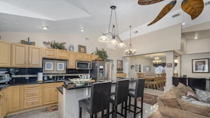 A spacious kitchen with wooden cabinets, a central island with bar stools, a dining area with a chandelier, and a living room with a sofa and pillows. - 10Bd/10Ba Large Beach Home, Indoor Pool In Virginia Beach (Virginia beach)