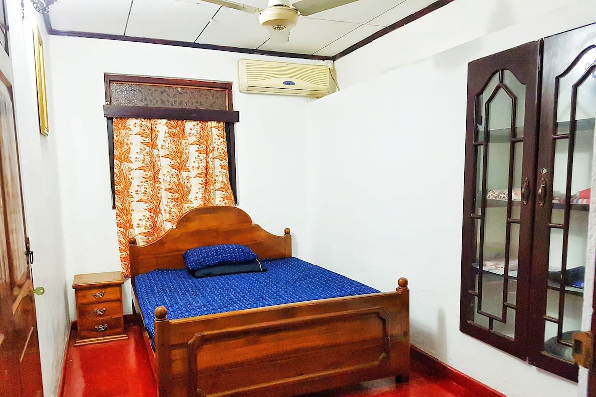 A bedroom with a wooden bed, a nightstand, a window with curtains, a ceiling fan, and a wall-mounted air conditioner.