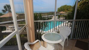 A balcony with a glass table and white wicker chairs, overlooking a pool and a beach.