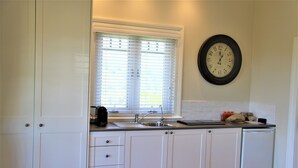 A kitchen with white cabinets, a sink, a window with blinds, and a wall clock. - Broughton Cottage - Pudding Hill Farm, Berry (Foxground)
