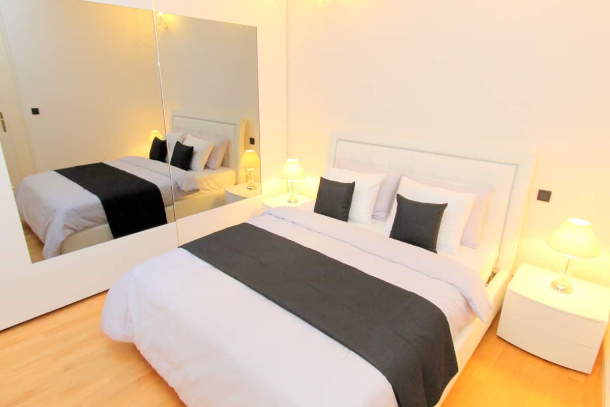 A neatly made bed with white and black bedding, a bedside table with a lamp, and a large mirror on the wall.