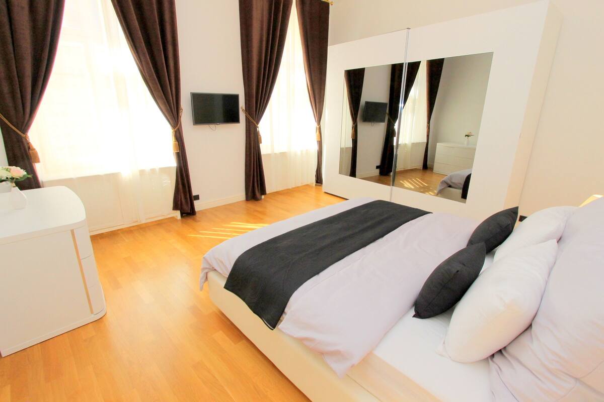 A bedroom with a bed, a bedside table, a mirror, and a window with curtains.