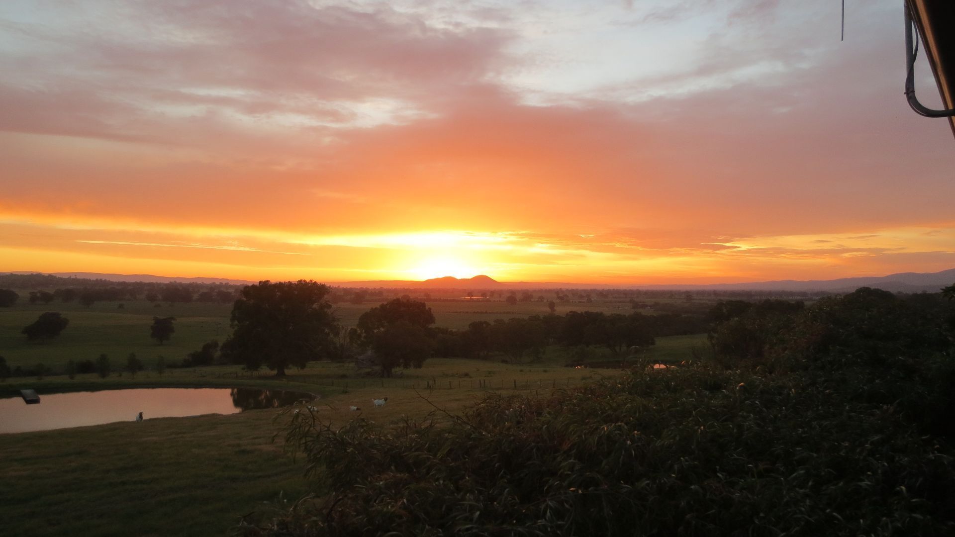 Ein Sonnenuntergang über einer ländlichen Landschaft mit Feldern, Bäumen und einem kleinen Gewässer.