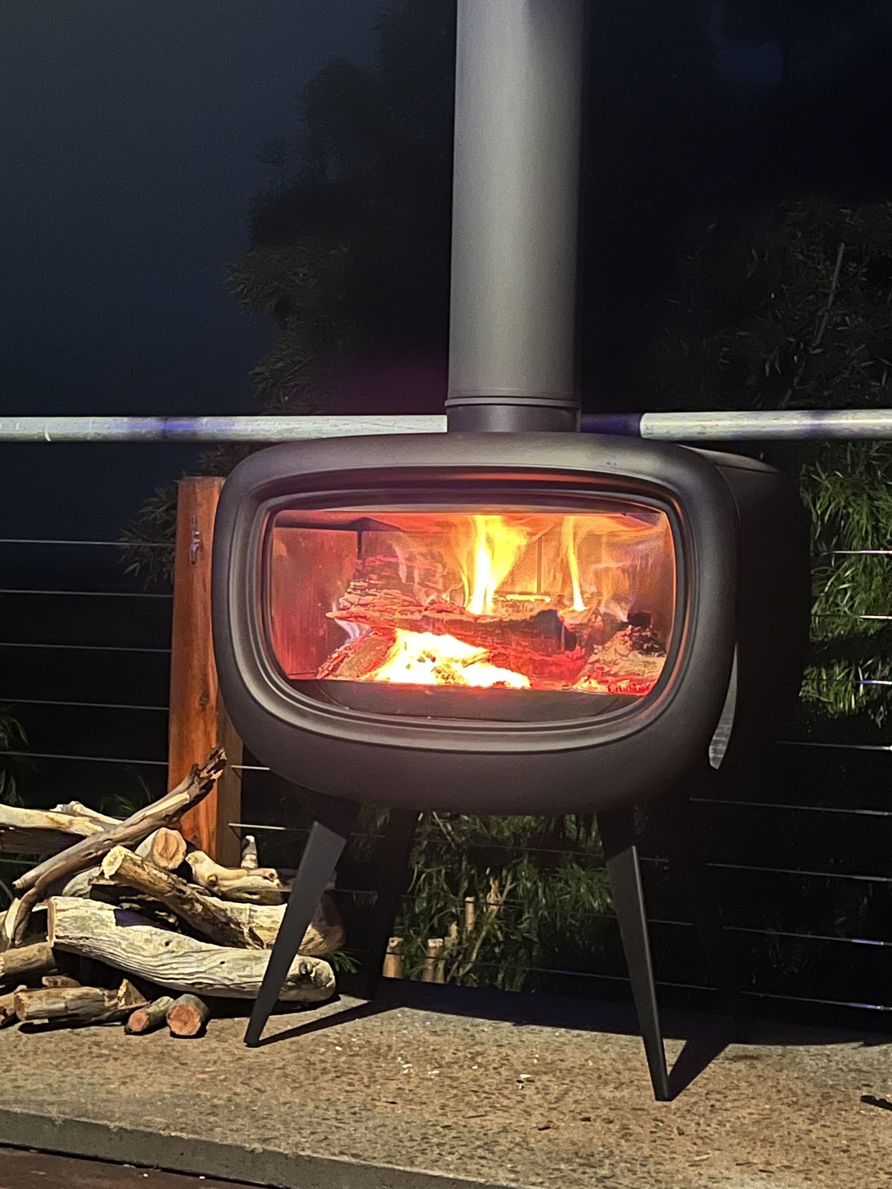 A wood-burning stove with a visible fire inside, placed on a stand with logs nearby.