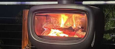 A wood-burning stove with a visible fire inside, placed on a stand with logs nearby.