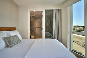 A hotel room with a large bed, a bathroom with a shower, and a view of the street through a large window. - Mayumi (Culver City)